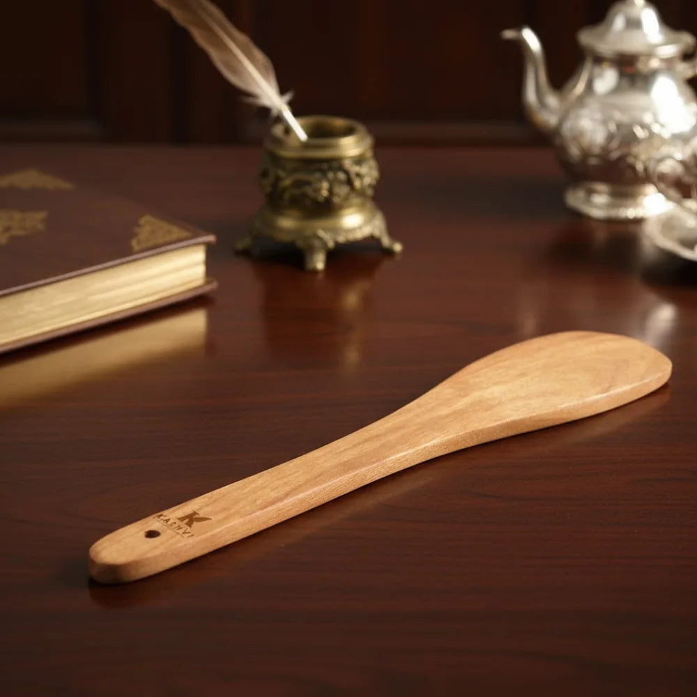 Wooden spoon on a wooden surface with a book, inkwell, and teapot in the background.